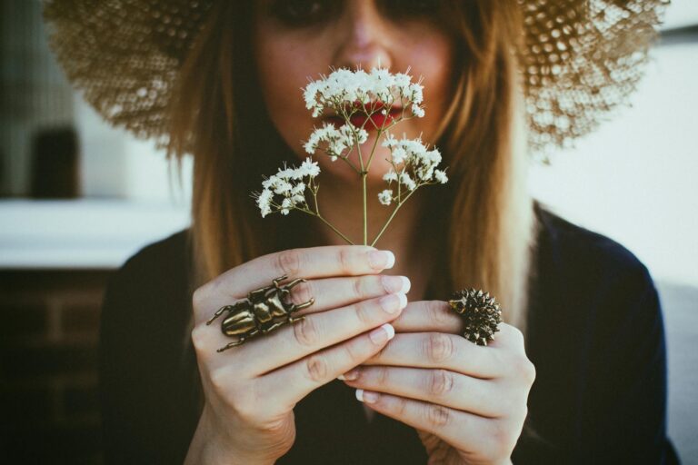 A woman in a straw hat holding delicate white flowers, showcasing unique beetle and spiked rings on her fingers.
