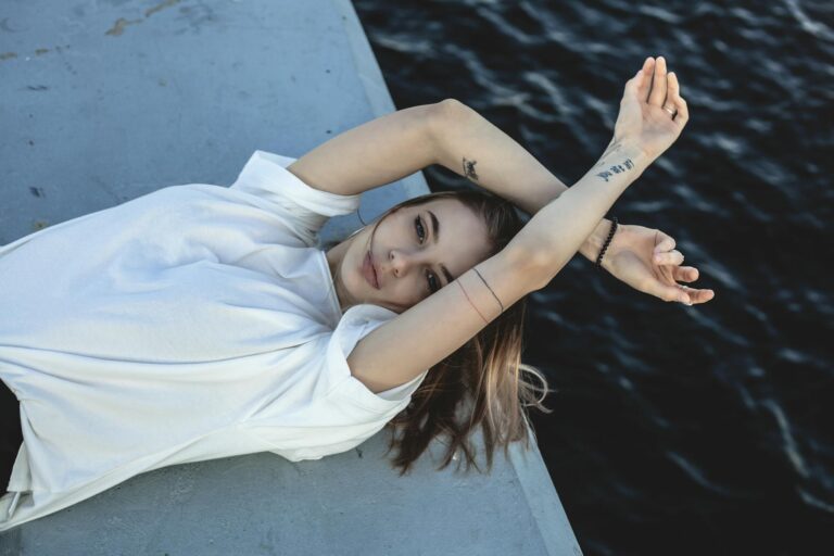 A young woman in a white t-shirt poses artistically by the ocean, exuding a sense of relaxation.