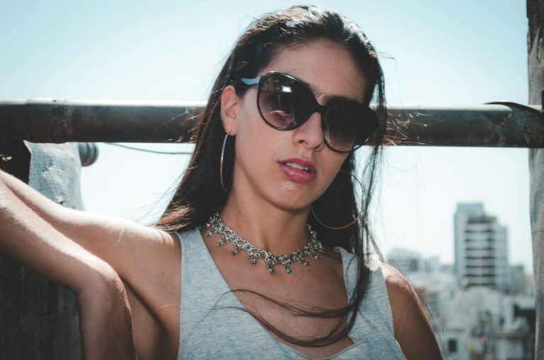 Elegant woman posing outdoors wearing sunglasses and a necklace against a city skyline.